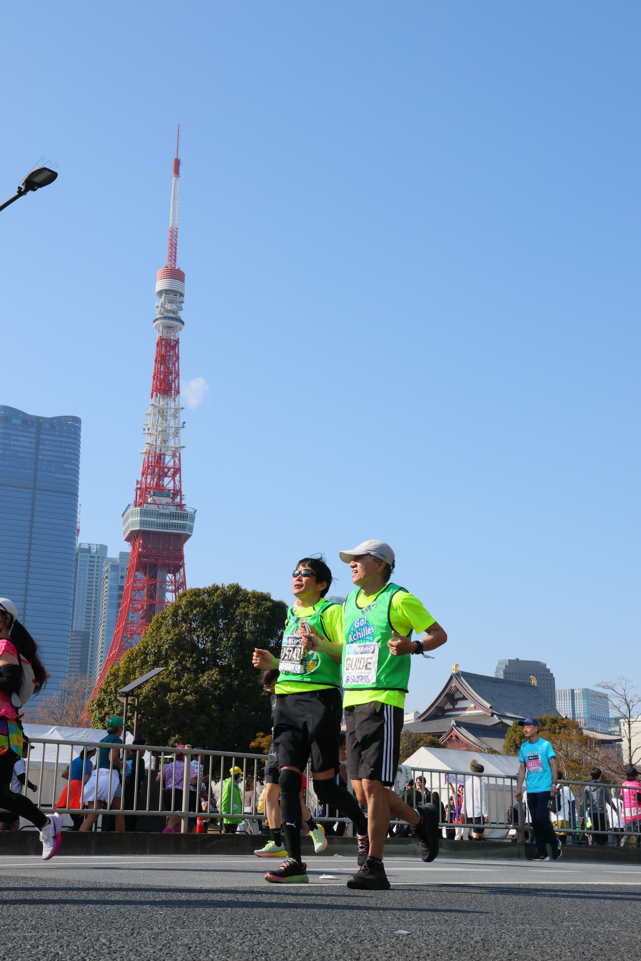 画像には、マラソン大会の様子が写っています。  手前には、ランナーたちが道路を走っている様子が捉えられています。男性2人が並んで走っており、蛍光色のグリ.jpg